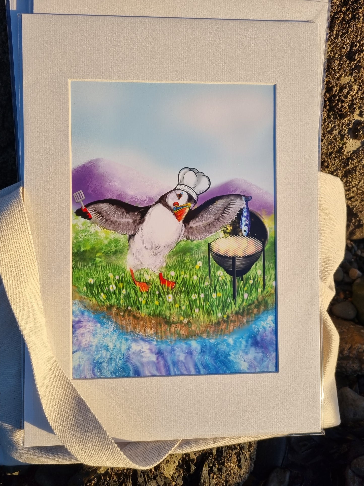 Whimsical puffin illustration of a puffin barbecuing a fish, created in mixed media with watercolour, gouache, and digital art. Illustrated on Anglesey by Welsh Coastal Artist Rachel Bennett.