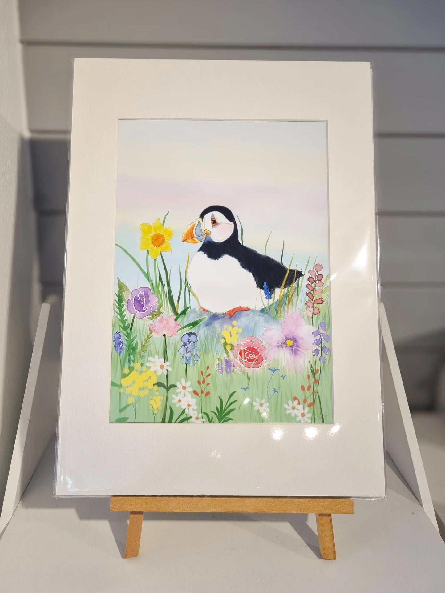 A hand-illustrated puffin, sitting amongst Spring Flowers, featuring a Welsh Daffodil. Painted on Anglesey by Welsh coastal/puffin artist Rachel Bennett.