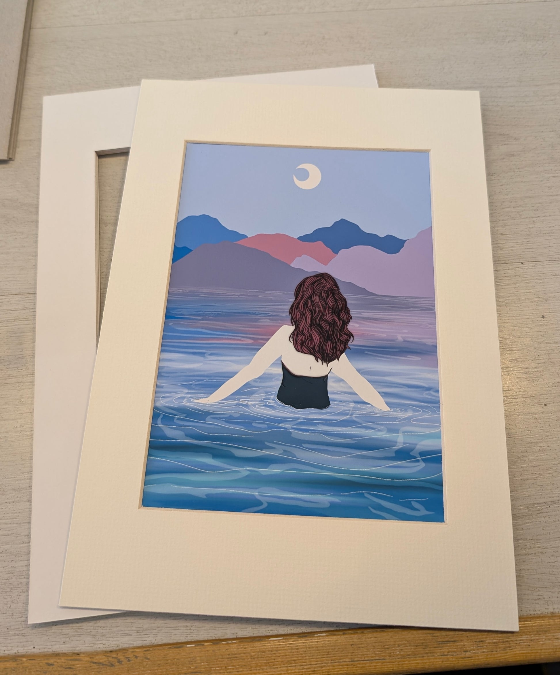 Illustration of a person about to swim in a lake with mountains at sunset, framed in a white mat on a wooden surface.